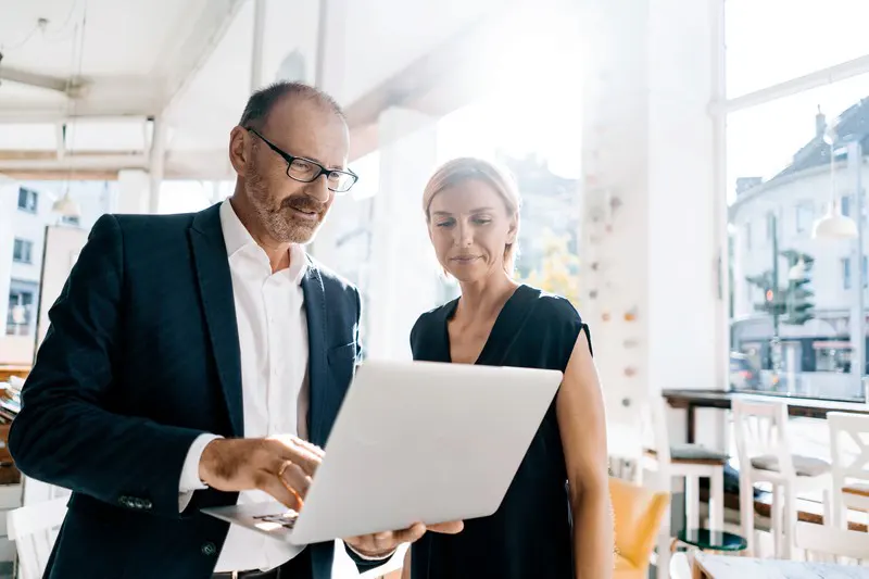 Two real estate agents, a man in a suit and a woman in black, review listings on a laptop in a bright, modern office.