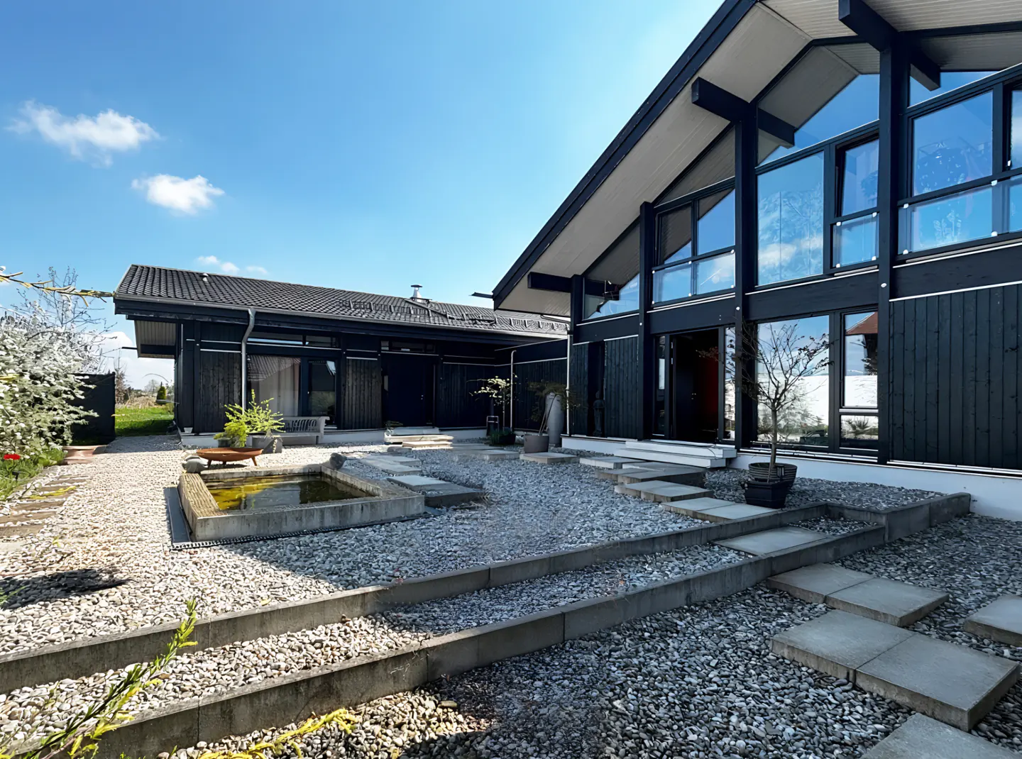 Exterior of a modern black house with large windows, a gravel yard, and a small pond.