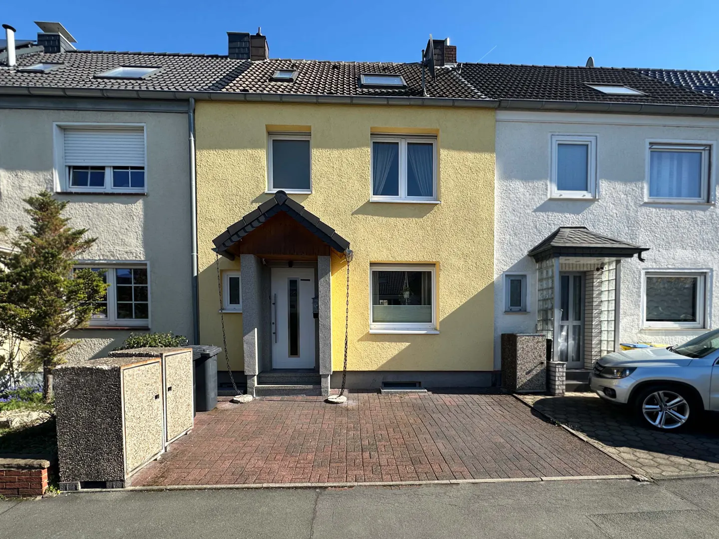 A yellow two-story townhouse with a dark roof and a small porch. The house is flanked by two similar townhouses.