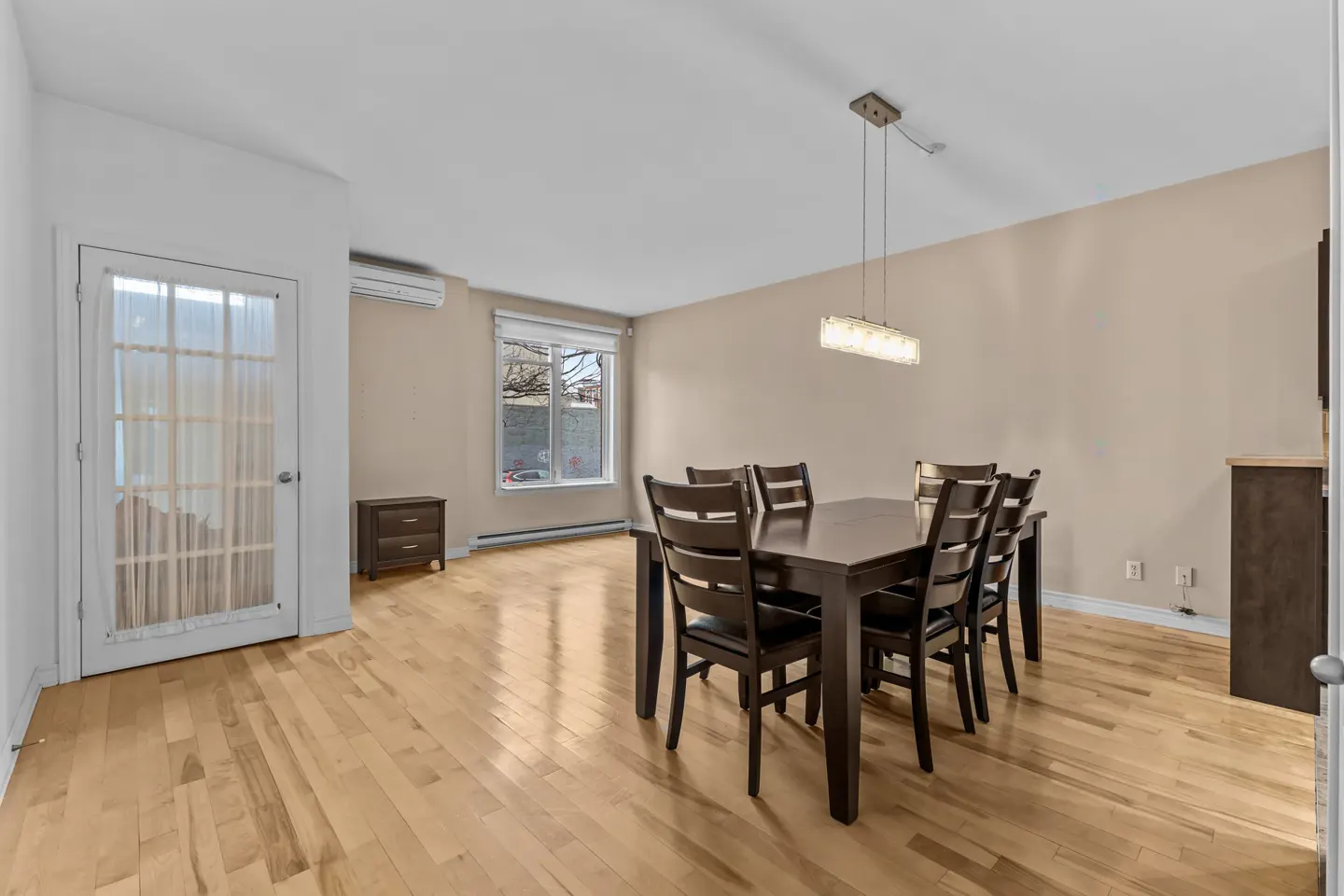 A dining room with a dark wood table and chairs under a modern light fixture. A window and door let in natural light.