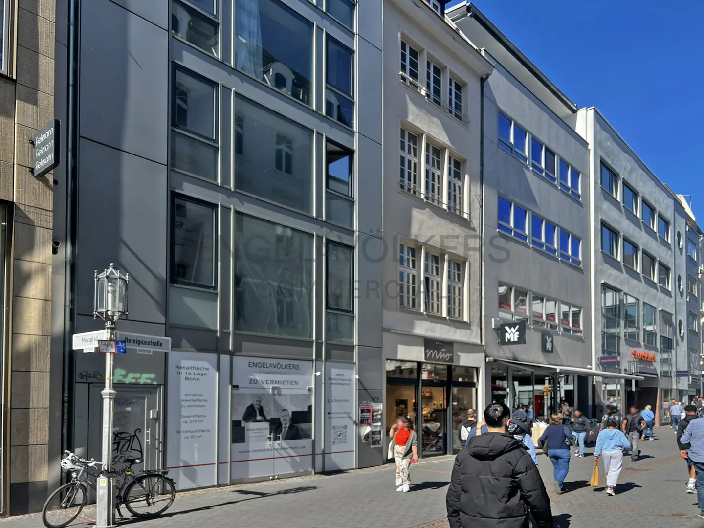 Street view of commercial buildings with shops and Engel & Völkers real estate signage. People walk along the sidewalk. Bicycles parked near a street lamp.