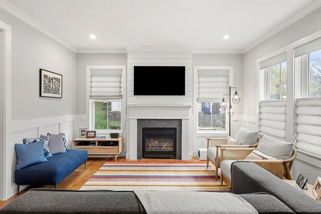Living room with a fireplace, TV, blue sofa, and two chairs. A colorful striped rug is on the floor. Windows with white blinds are on either side of the fireplace.