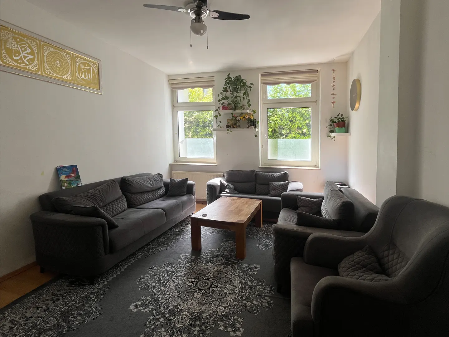 Living room with gray sofas, a wooden coffee table, and a patterned rug. Two windows let in natural light. A ceiling fan is visible.