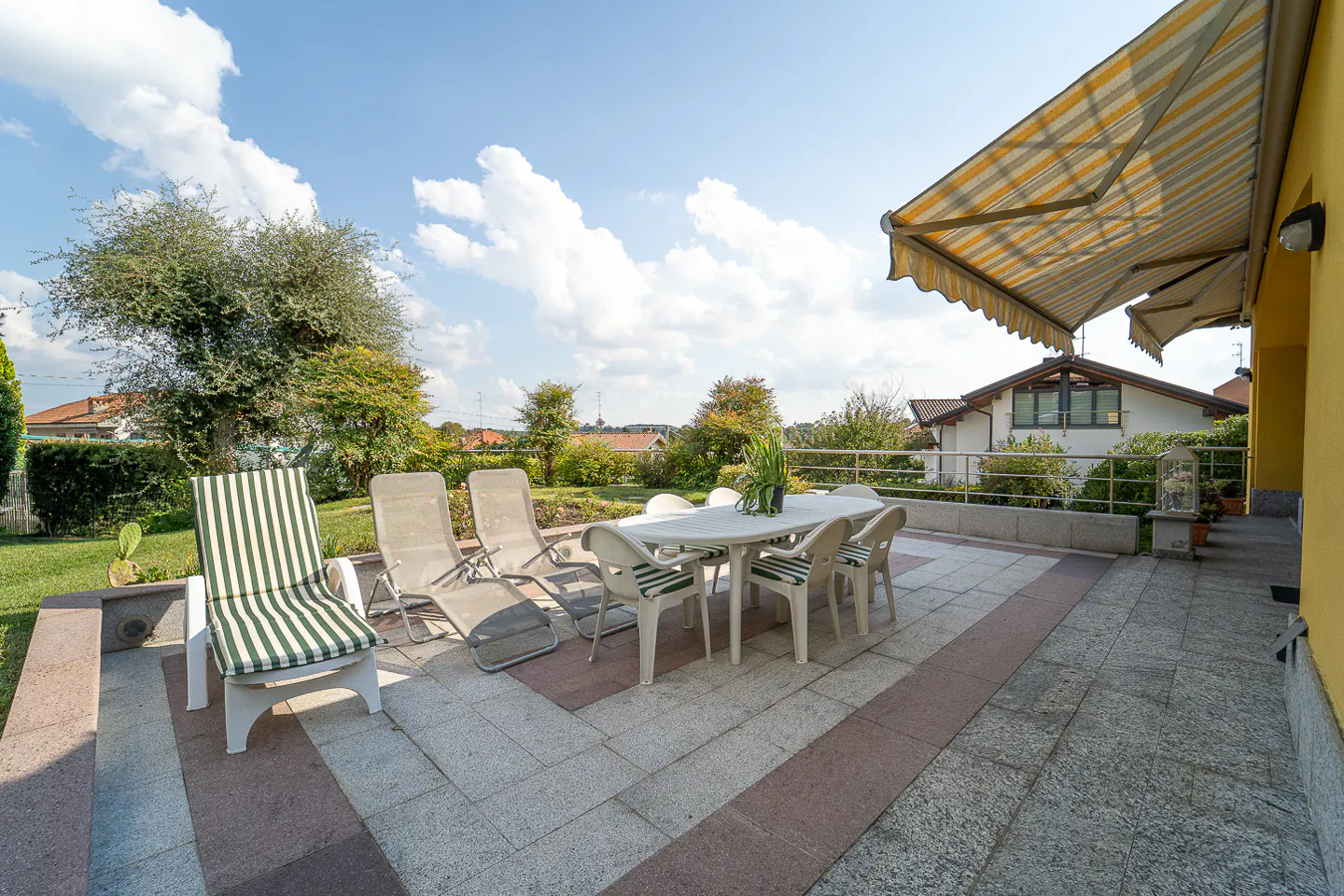 Outdoor patio with a white table, chairs, lounge chairs, and a striped awning. Green lawn and trees in the background.