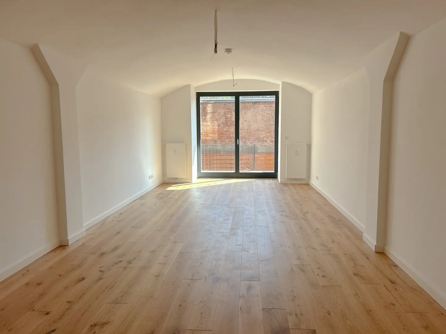 Bright, empty room with light wood floors and white walls. A black-framed sliding glass door leads to a brick building.