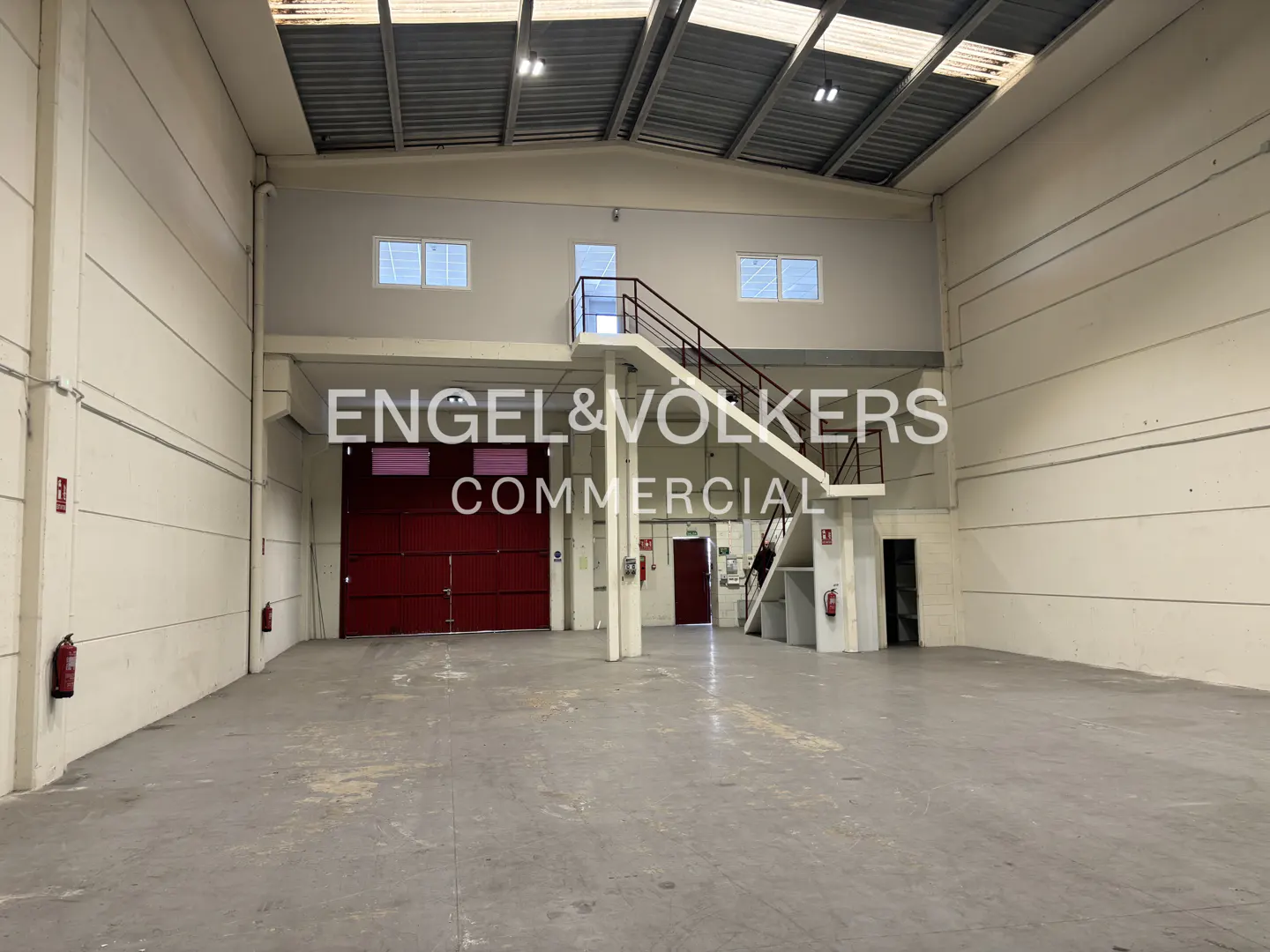 A large, empty warehouse with a red door and a metal staircase. The walls are white and the floor is gray. There are windows on the upper level.