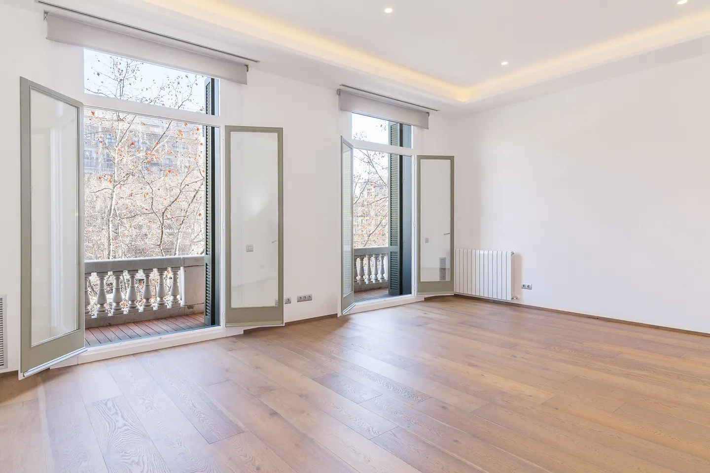 Bright, empty room with light wood floors, white walls, and two sets of open balcony doors overlooking trees.