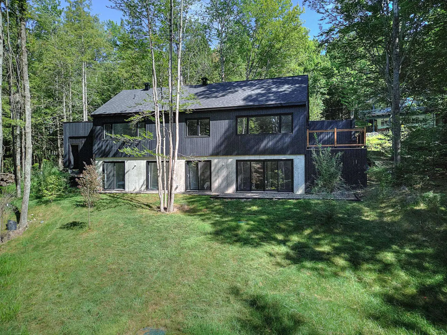 Two-story modern home with black siding and a concrete foundation, surrounded by green trees and a grassy lawn.