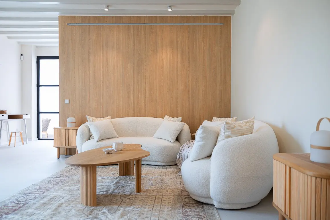 Bright living room with white walls, a wood accent wall, and white furniture. A patterned rug sits under a wood coffee table.
