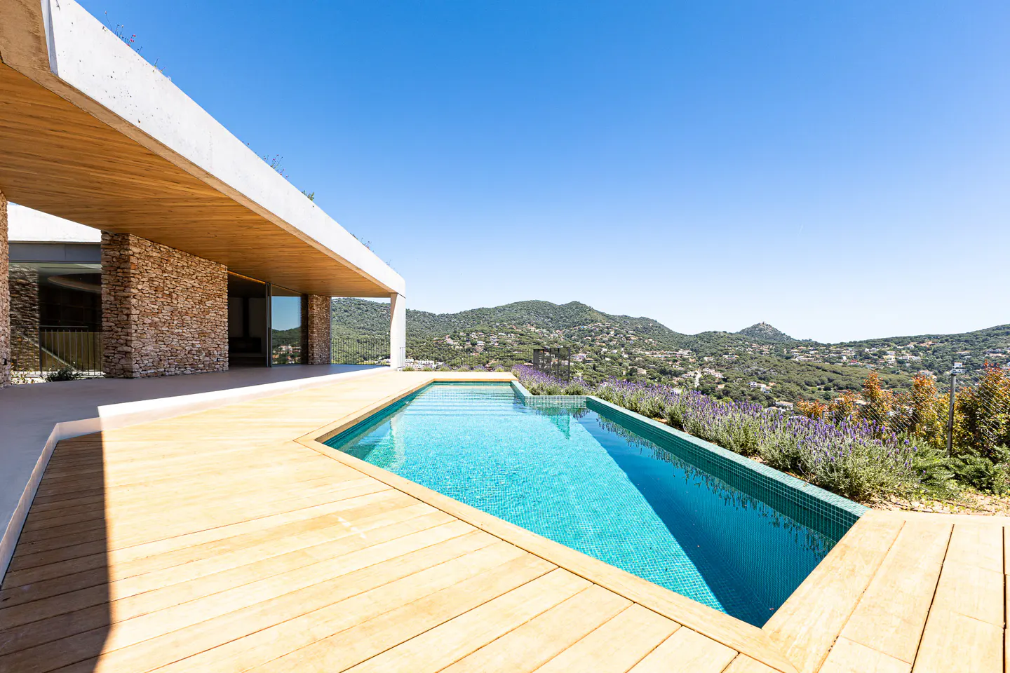 Modern home with a blue tiled pool and wood deck overlooking a green hillside under a clear blue sky.