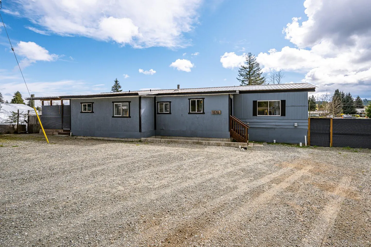 A single-story, light blue house with a gravel driveway under a partly cloudy sky.