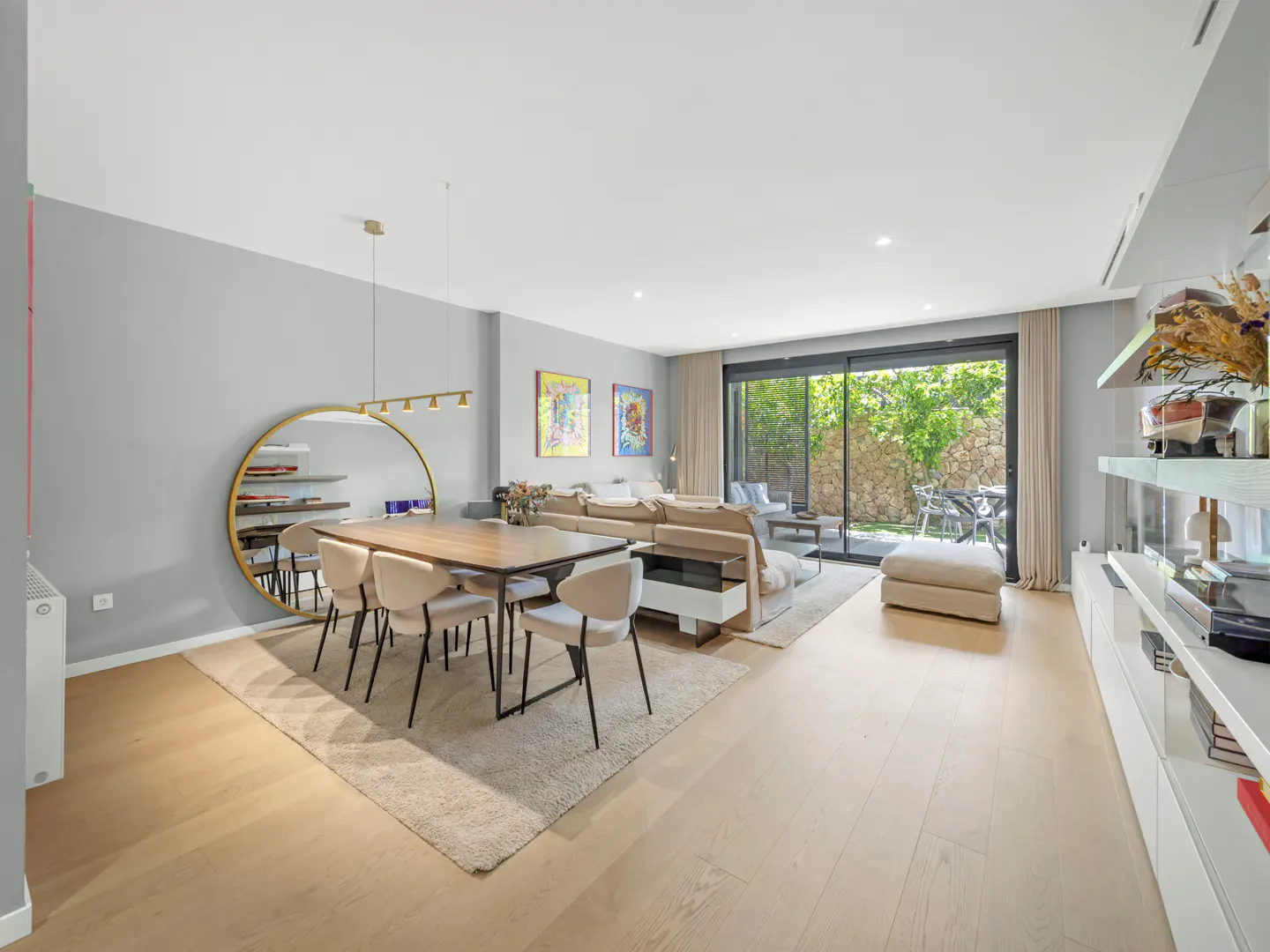 Bright, open-plan living and dining area with light wood floors. A large round mirror and dining table with chairs are on the left. Sliding glass doors lead to a patio.