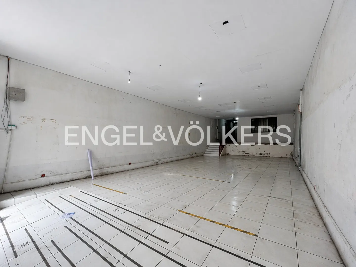 Empty commercial space with white tile floor, white walls, and overhead lighting. Engel & Völkers logo on the wall.
