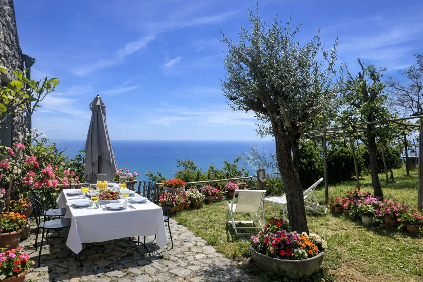 Outdoor dining area with ocean view. Table set with white tablecloth, plates, and drinks. Flowers in pots line the patio. Blue sky.