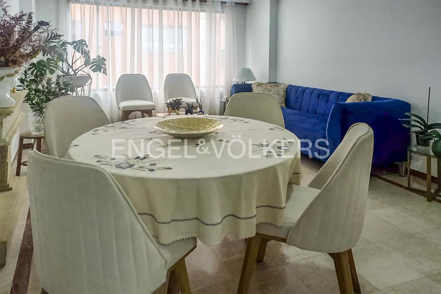 Living room with round table, six chairs, and a blue sofa. A golden bowl sits on the table. Engel & Völkers logo is visible.