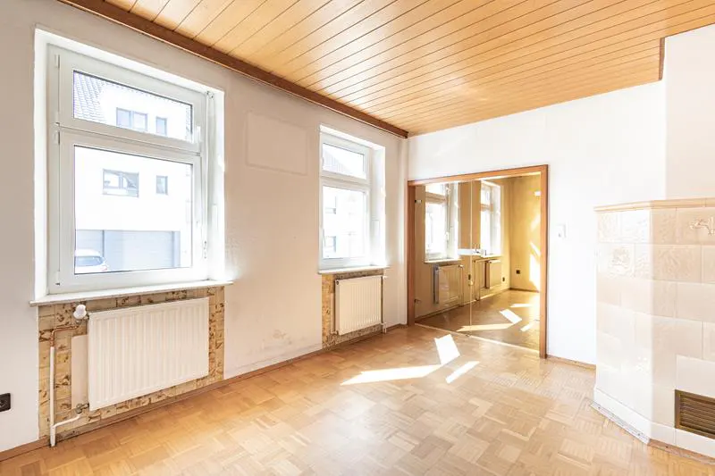 An empty room with white walls, wood floors, and a wood-paneled ceiling. Two windows let in natural light. A doorway leads to another room.