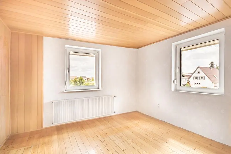 Bright, empty room with wood floors, ceiling, and partial wall. Two windows show a house and trees outside. A white radiator sits under one window.
