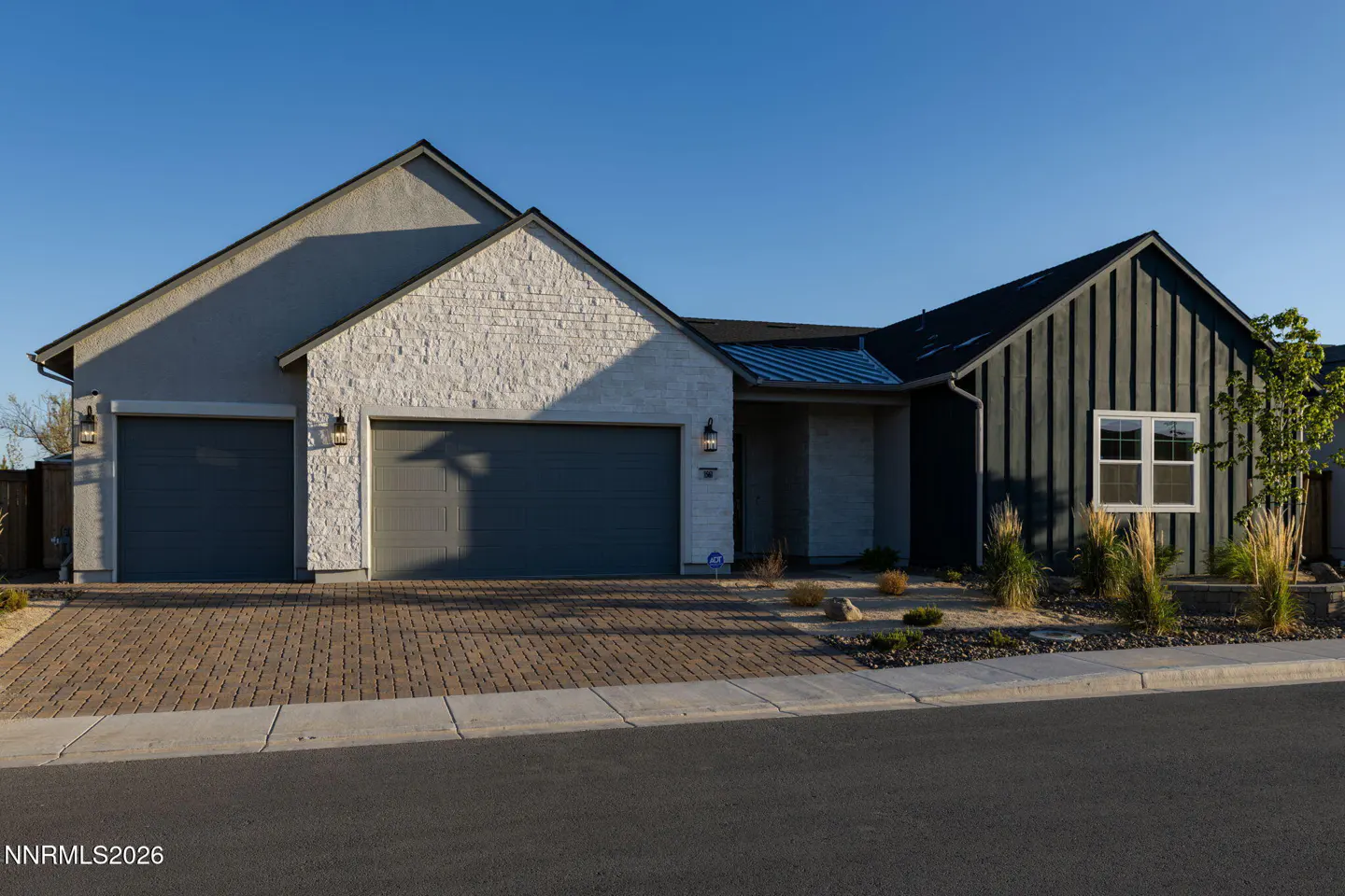 Exterior of a modern, single-story home with gray siding, stone accents, and a brick driveway under a clear blue sky.