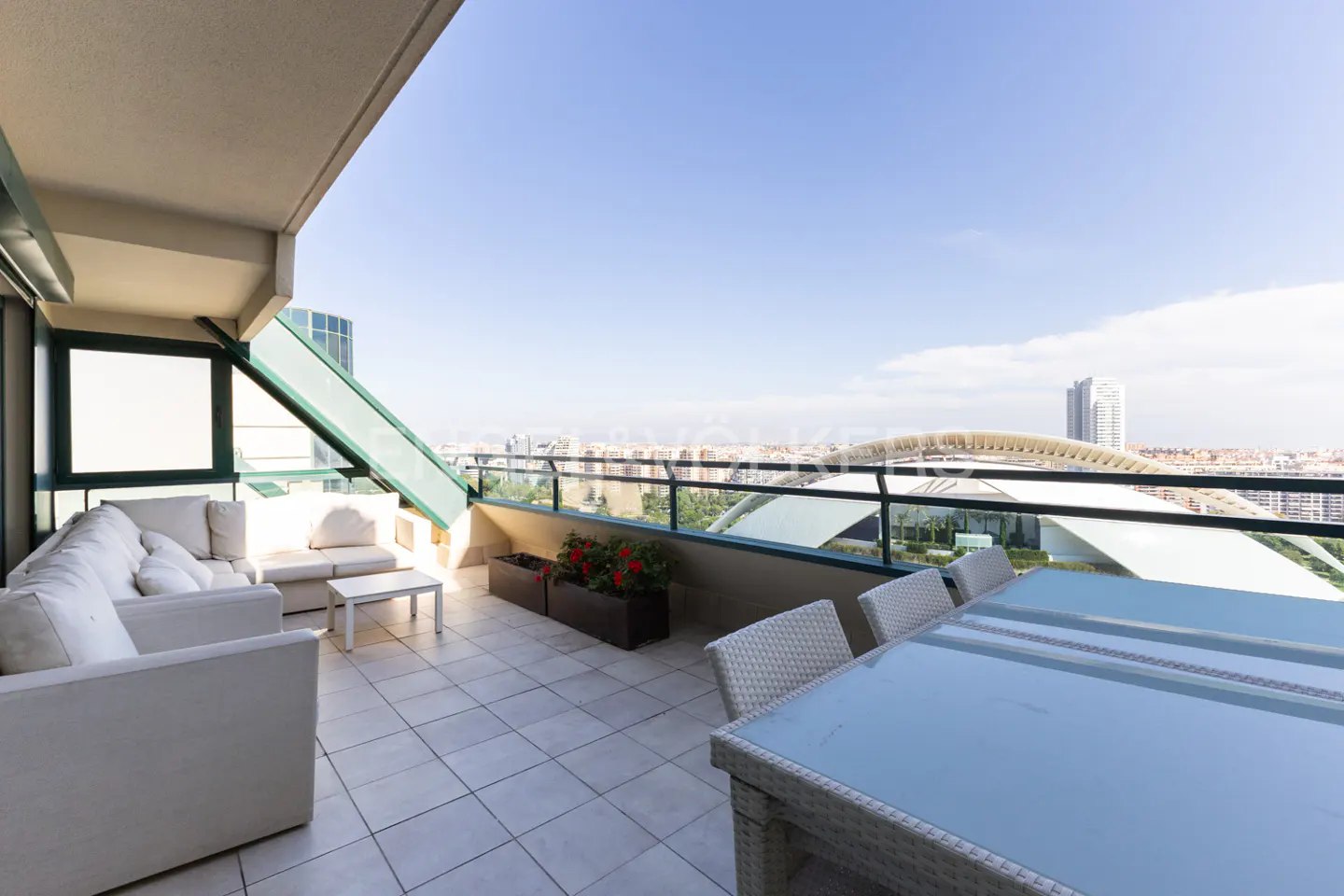 Balcony with white furniture, glass table, and city view. Red flowers in planters line the railing. Blue sky above.