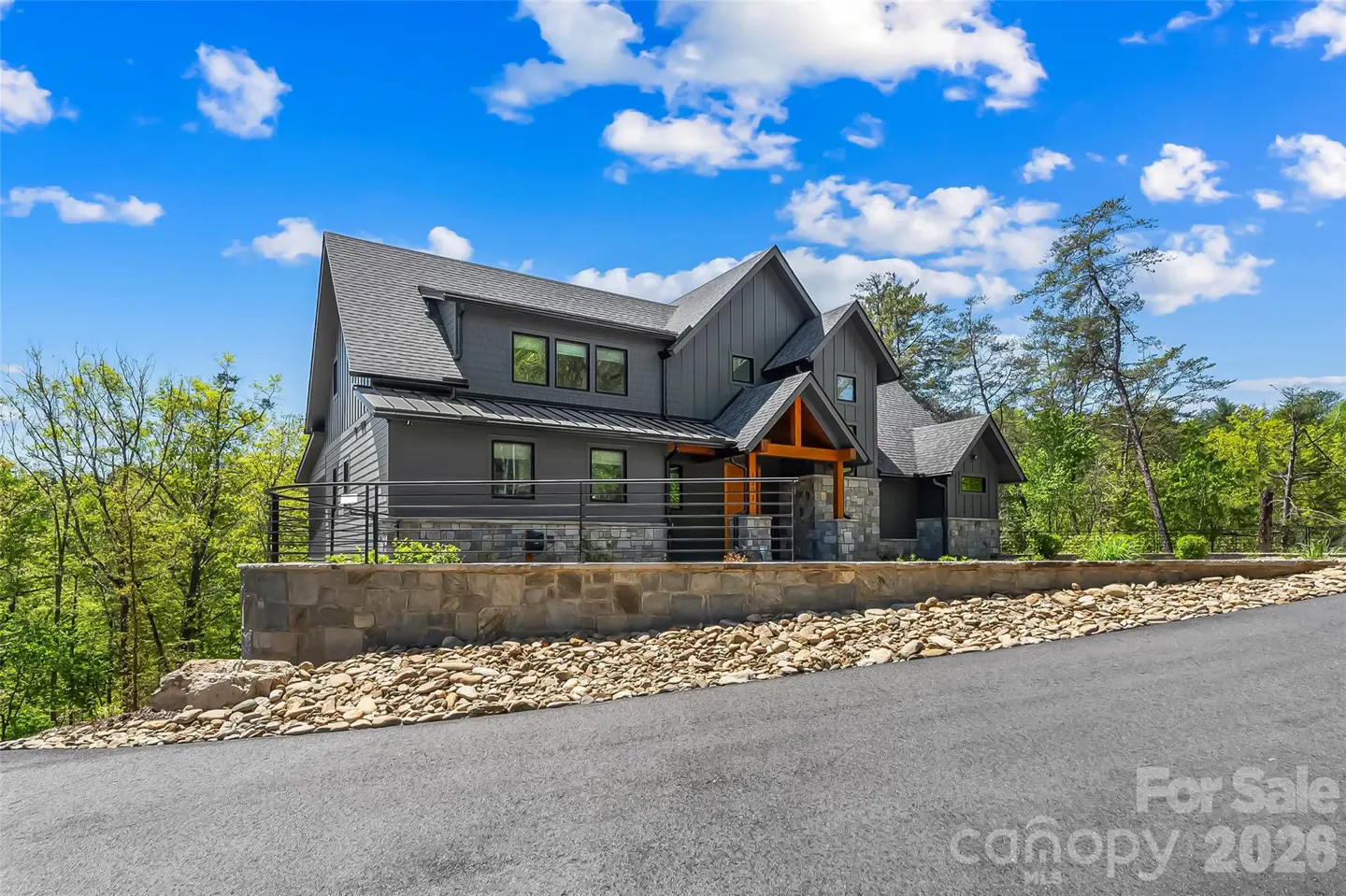 Modern gray house with wood accents, stone base, and black roof, set against a blue sky and green trees. "For Sale" text visible.
