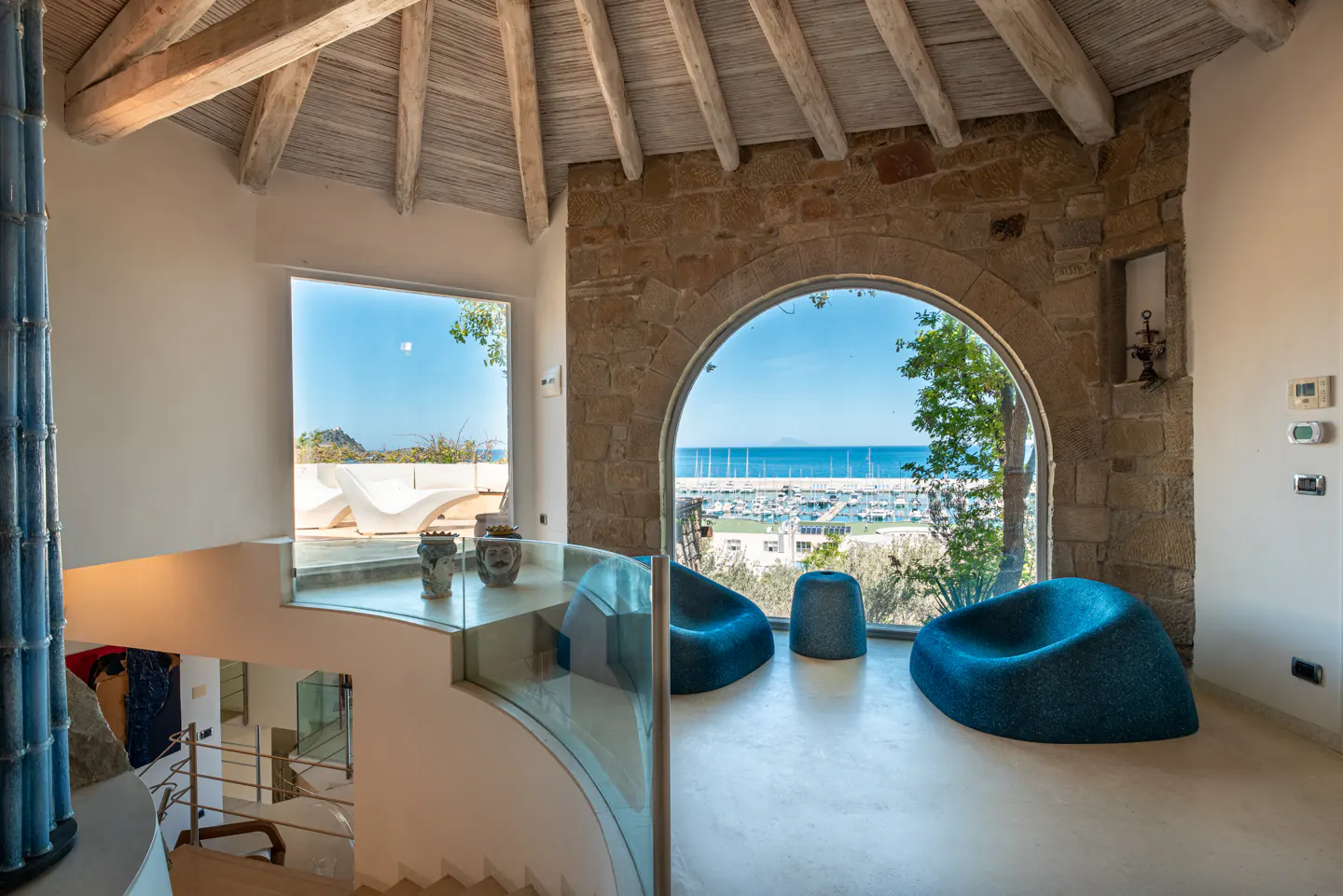 Interior view of a modern home with stone arch window overlooking a marina. Blue chairs sit on a white floor. A staircase is visible.