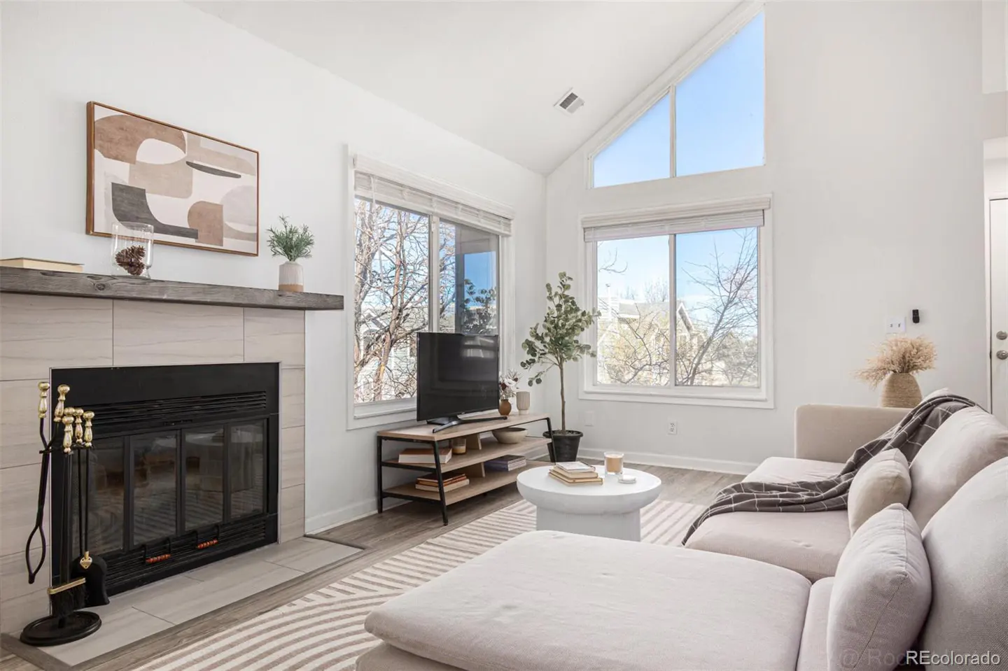 Bright living room with a fireplace, beige sectional sofa, and large windows. A TV sits on a wooden stand, and a round white table holds books.