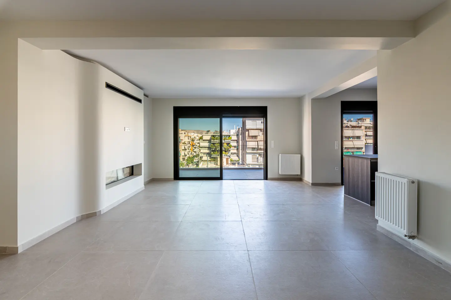 Bright, empty living room with gray tile floor, white walls, and black-framed sliding glass doors to a balcony with city views.