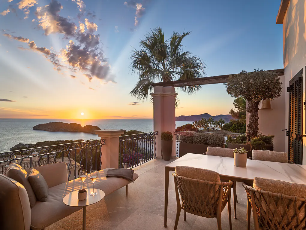 Balcony view at sunset. Lounge seating and dining table overlook the ocean and island. Palm tree and wrought iron railing.