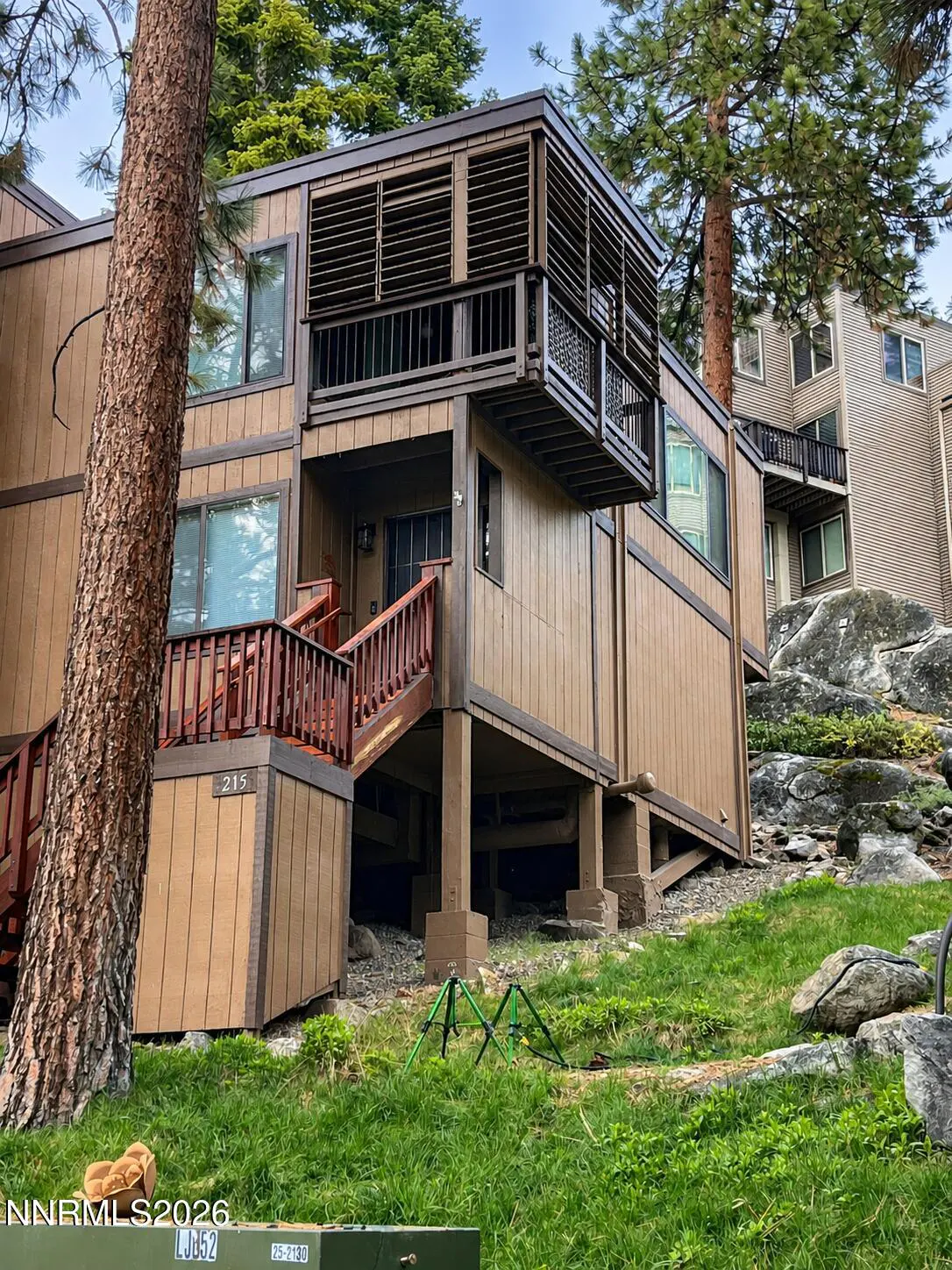 A two-story brown condo with a wooden deck and stairs, surrounded by trees and green grass.