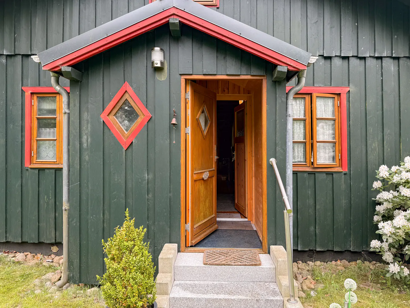 Exterior of a green wooden house with an open wooden door, red trim, and stone steps.