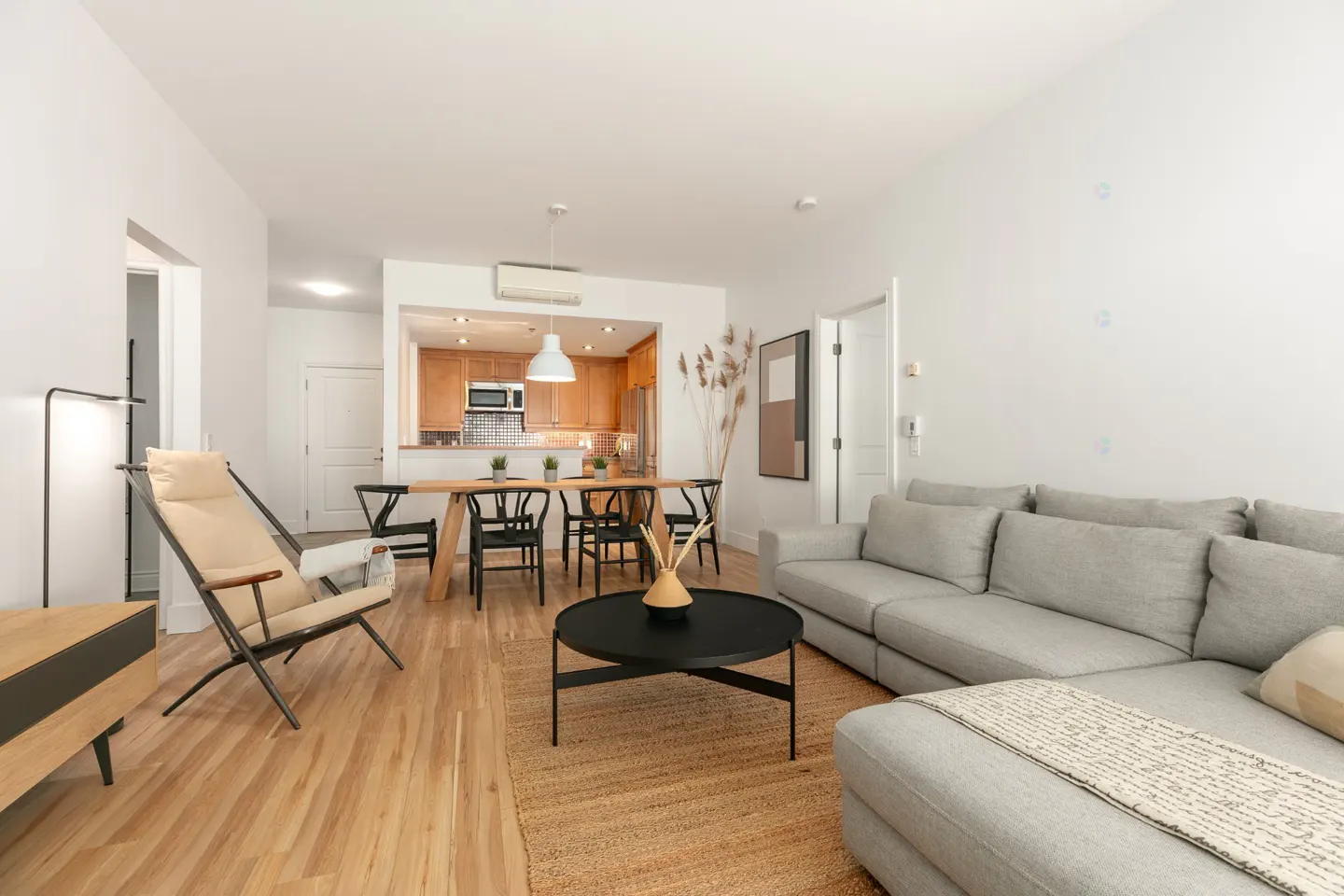 Bright, open-concept living space with a gray sectional sofa, black coffee table, and dining area with wooden table and black chairs. Kitchen visible in the background.