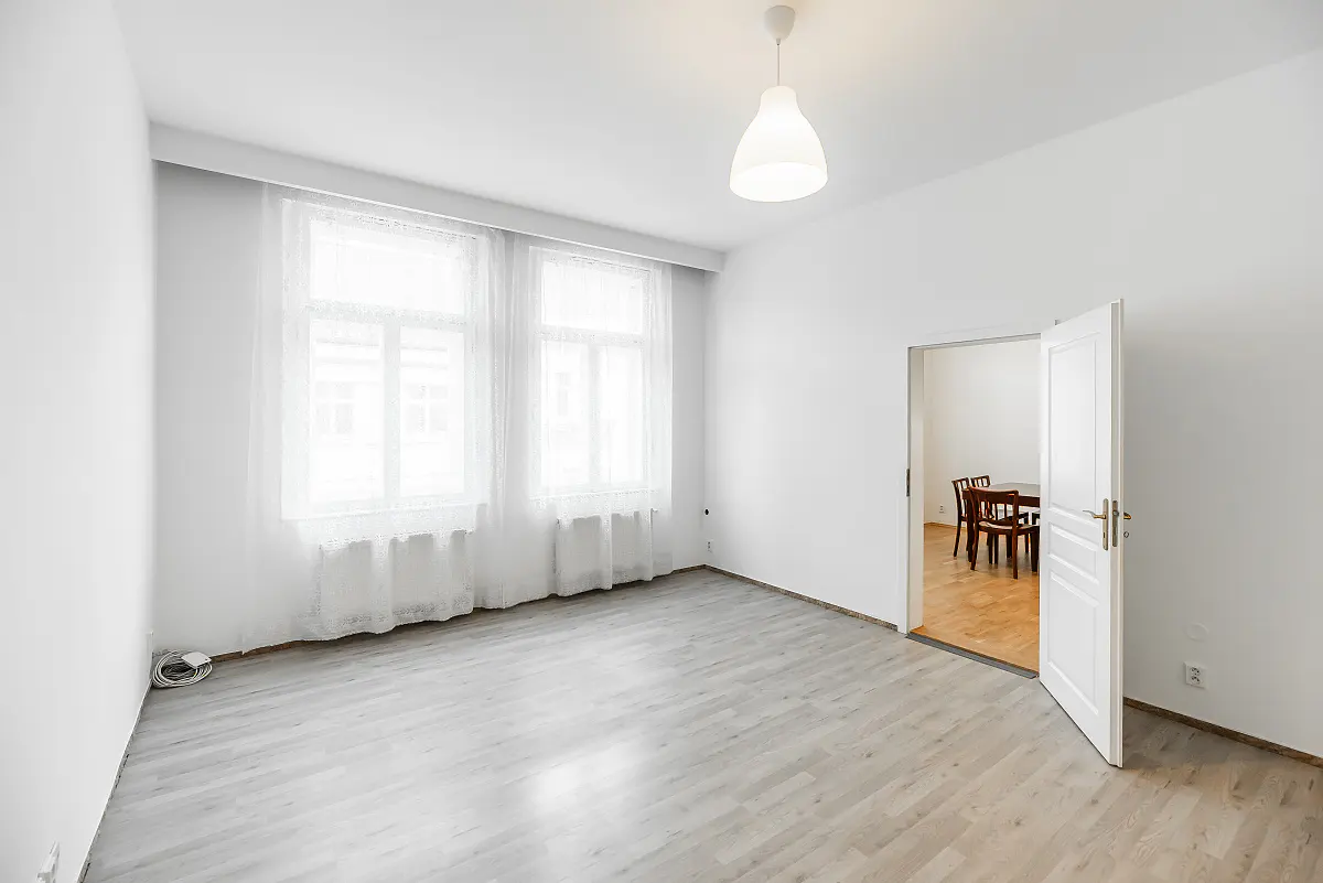 Bright, empty room with light wood floors, white walls, and two windows with sheer curtains. An open door reveals a dining table and chairs in the next room.