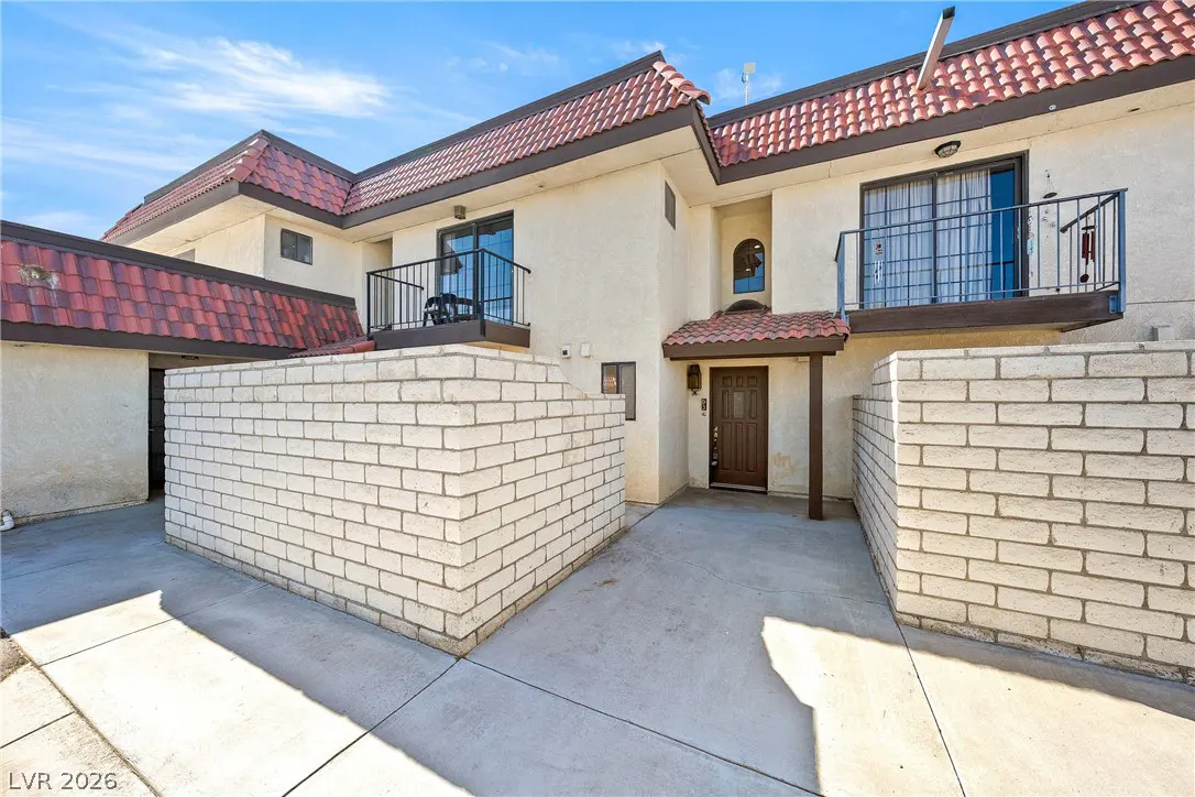 Exterior view of a two-story townhouse with a red tile roof, beige stucco walls, and small balconies with black railings. A brick wall encloses the front patio.