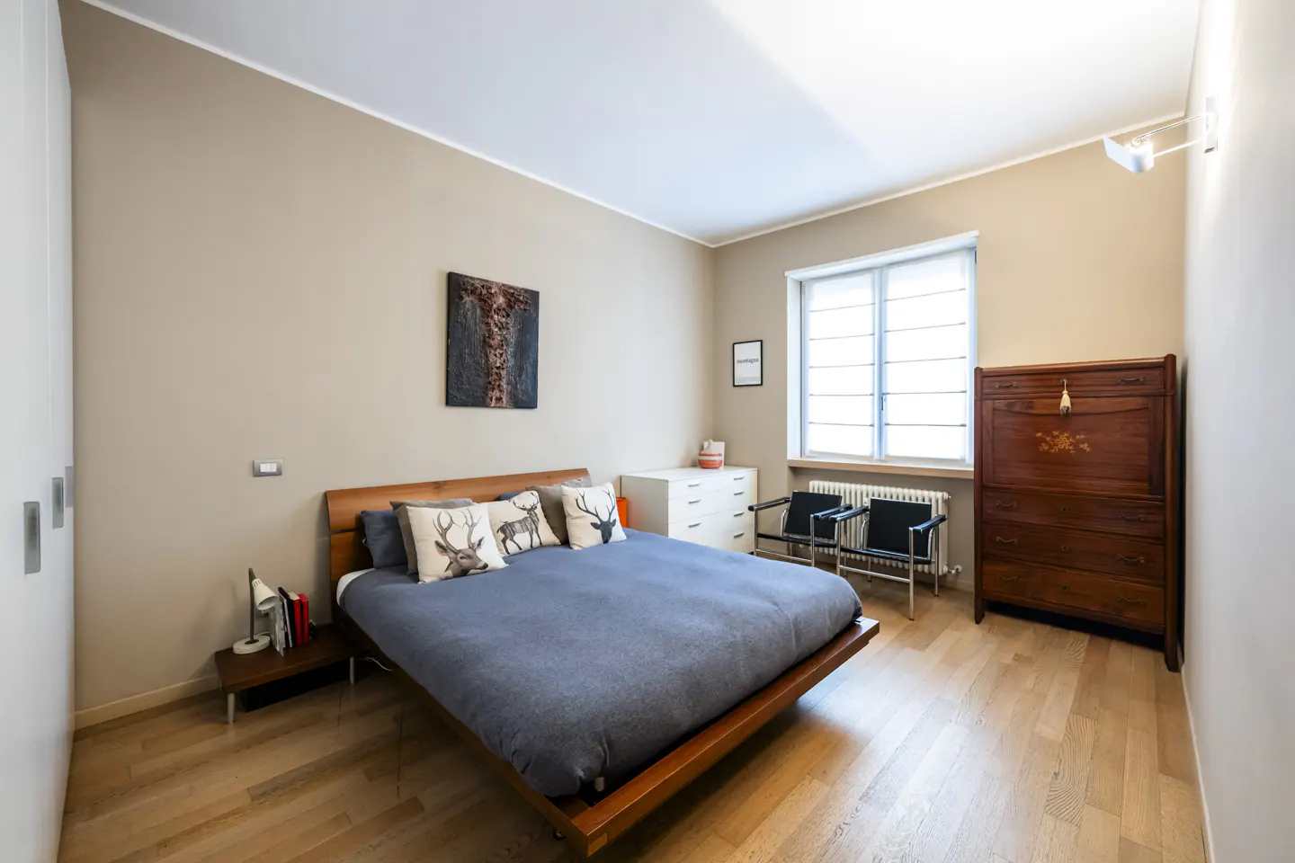 Bedroom with a wooden bed, blue duvet, deer pillows, and a dark wood antique dresser. Light wood floors and beige walls.