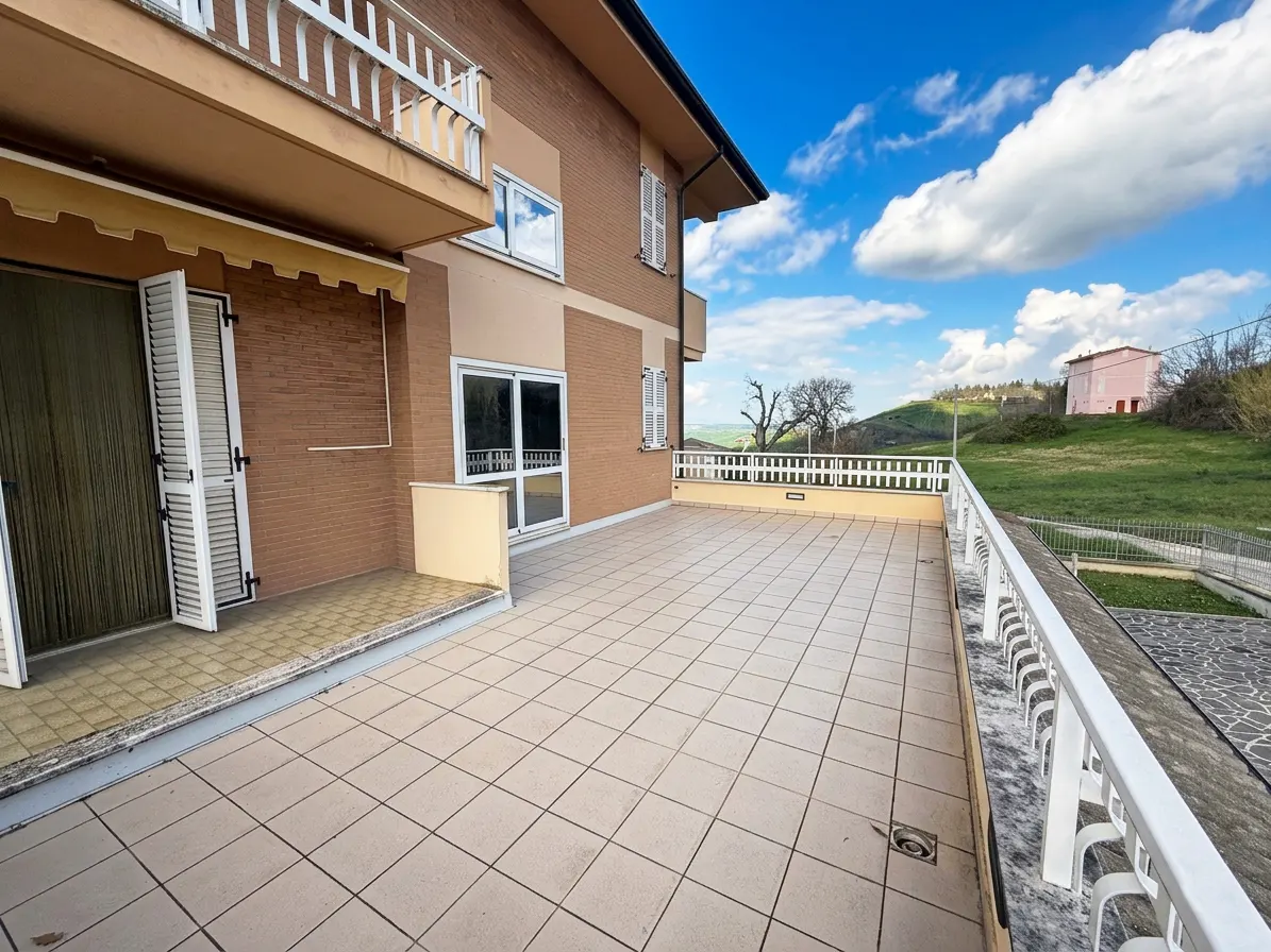 A large, tiled patio with a white railing overlooks a green, rolling landscape under a blue sky with fluffy clouds. The patio is attached to a two-story brick building.
