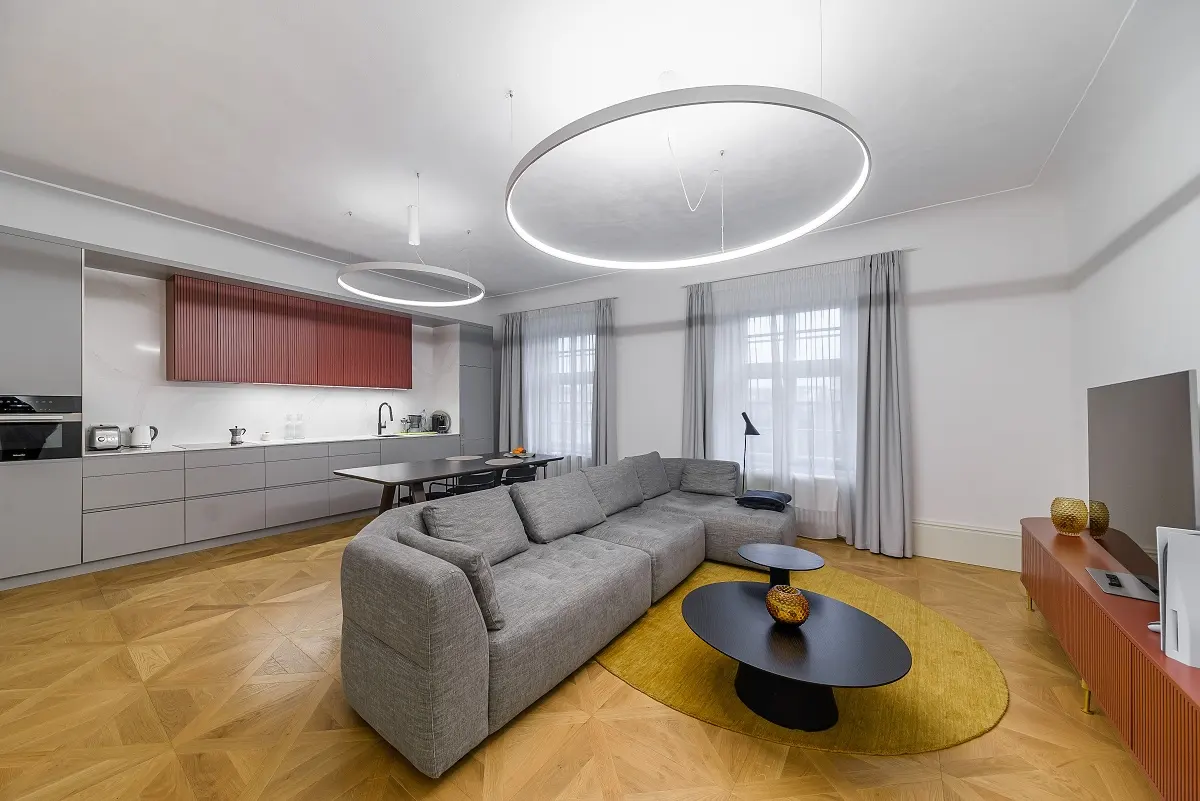 A modern living room with a gray sectional sofa, black coffee tables on a yellow rug, and a kitchen in the background.
