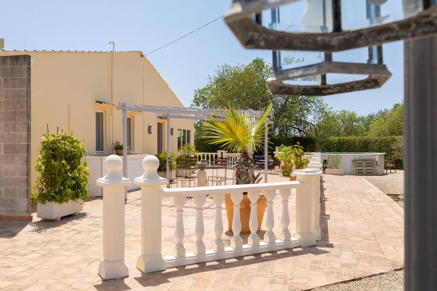 Exterior view of a cream-colored house with a brick patio, white balustrade, and a small palm tree. A metal pergola is attached to the house.