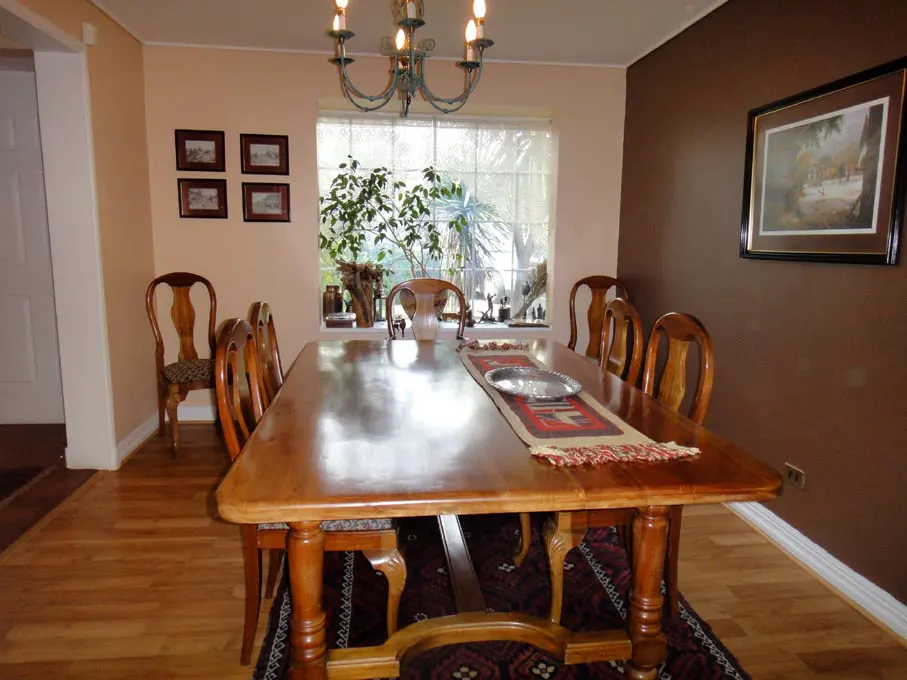Dining room with a long wooden table, chairs, and a chandelier. A window with plants is in the background. The walls are painted in brown and peach colors.