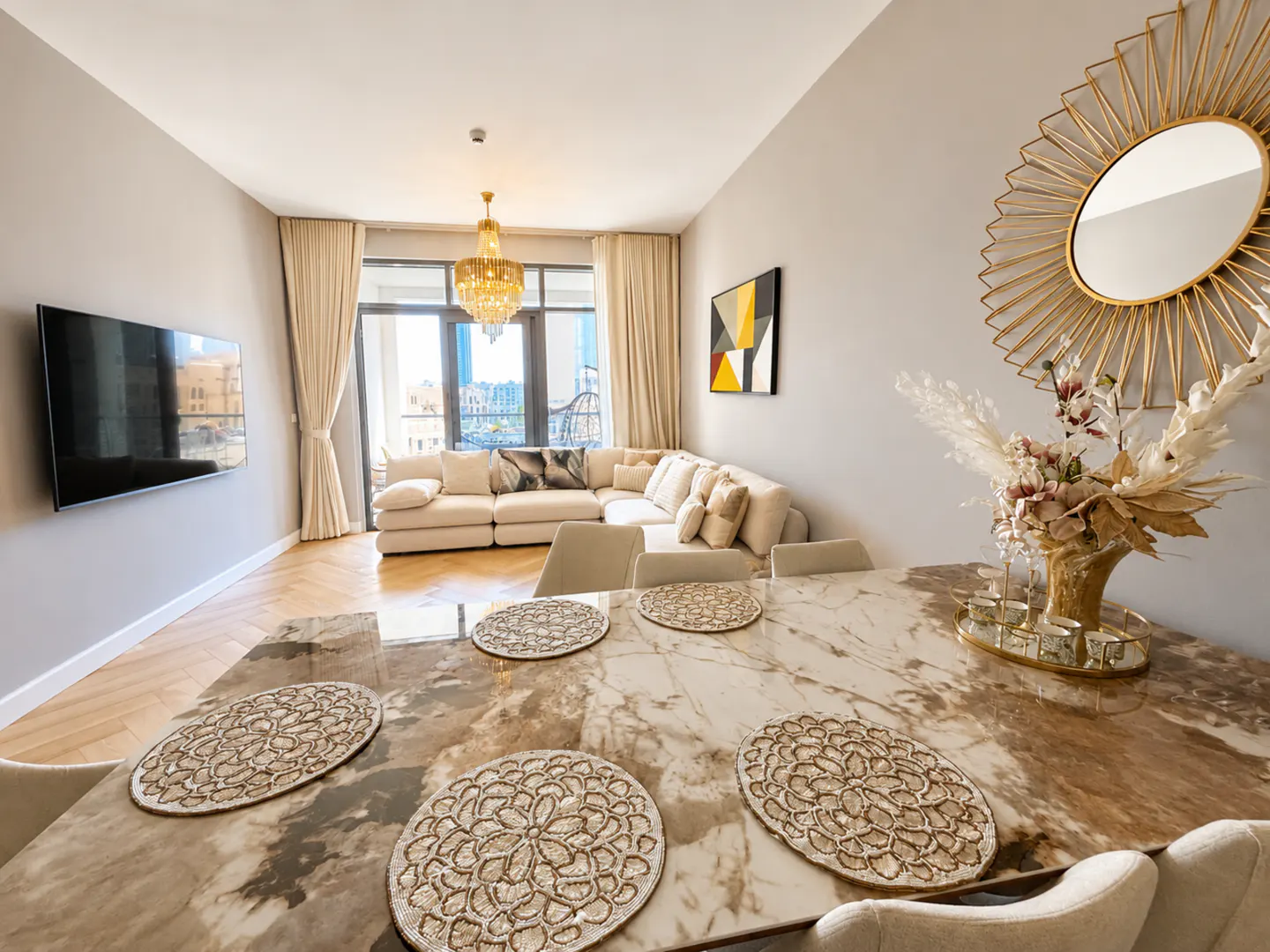 Modern living room with a marble dining table, beige chairs, a large sectional sofa, wall-mounted TV, and gold accents.