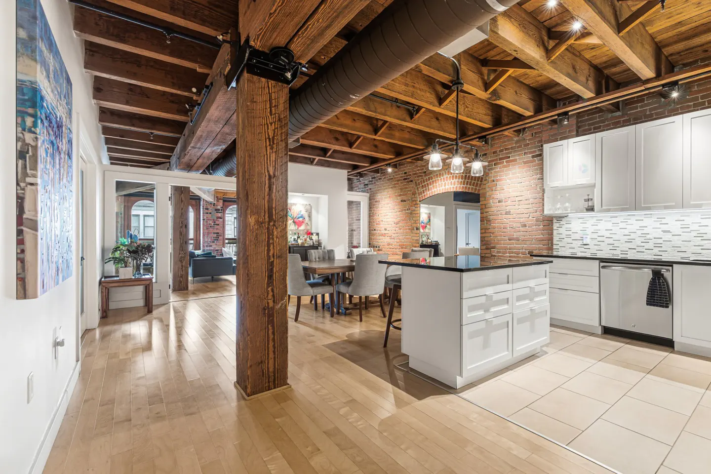 Open-concept loft with exposed brick, wood beams, and a white kitchen island. Hardwood floors and a dining area are visible.