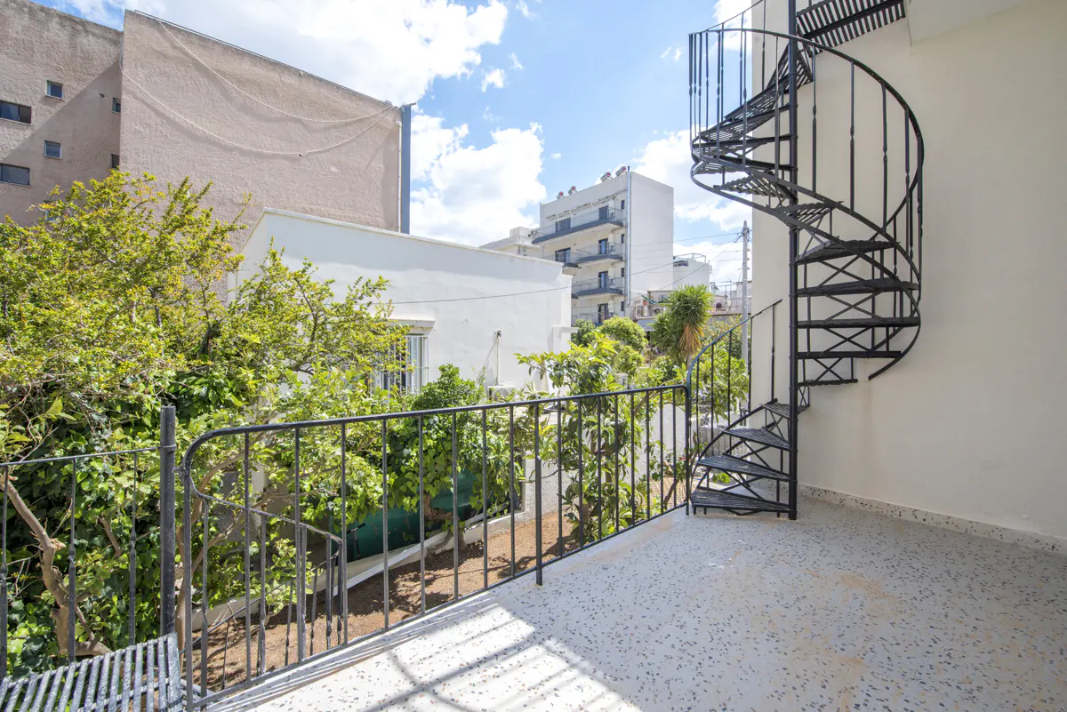 Balcony view with black spiral staircase, metal railing, and speckled floor. Lush green trees and buildings in the background under a blue sky.