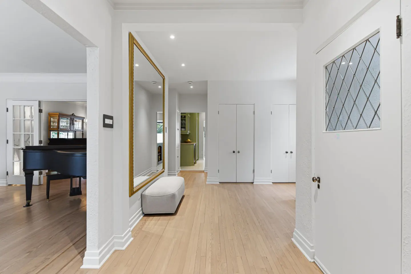 Bright hallway with light wood floors, white walls, and a large gold-framed mirror. A piano is visible through an open doorway.