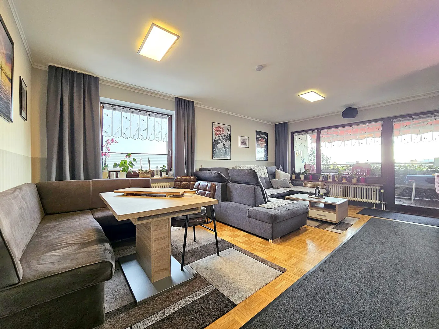 A living room with a gray sectional sofa, a dining table with chairs, and a balcony with plants.