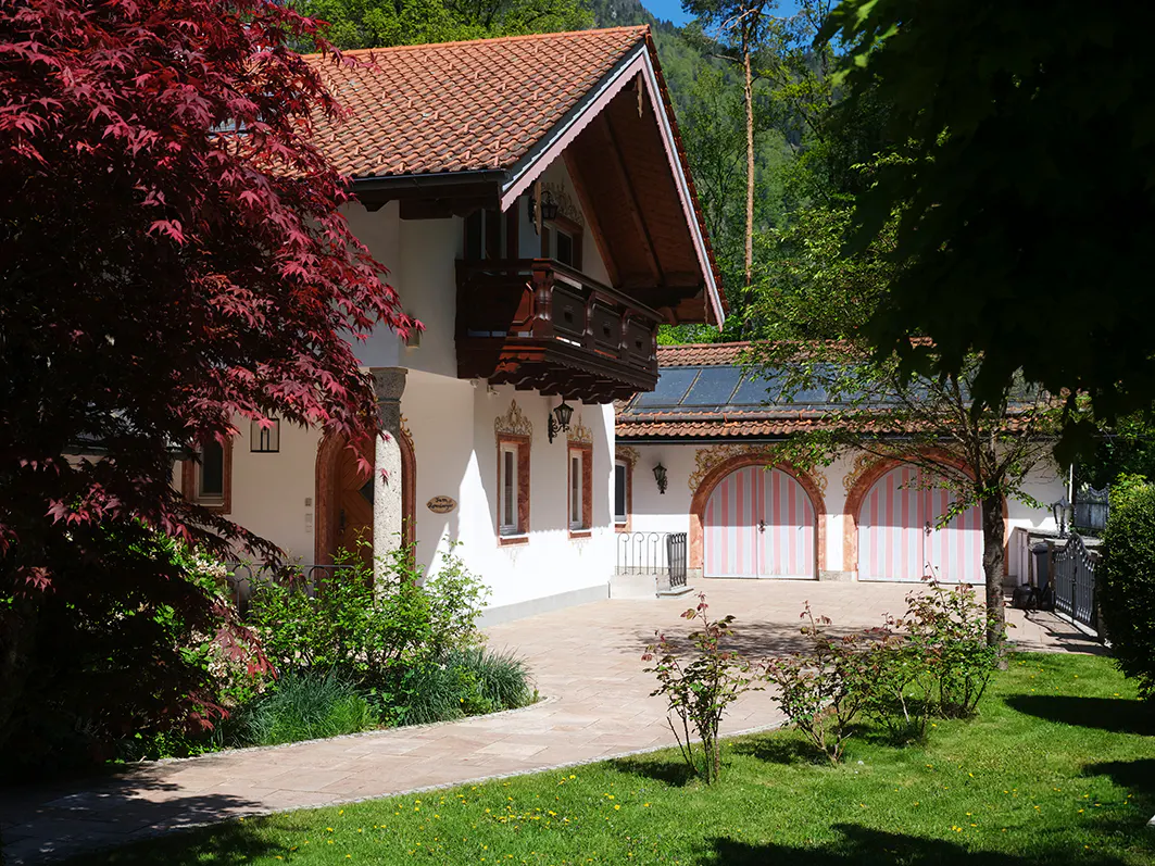 A white house with a red tile roof and a brown balcony, surrounded by green trees and grass.