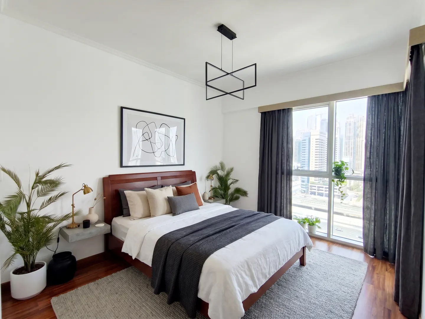 Bedroom with a wood bed frame, white bedding, gray throw, and pillows. A modern light fixture hangs above, and a city view is visible through the window.
