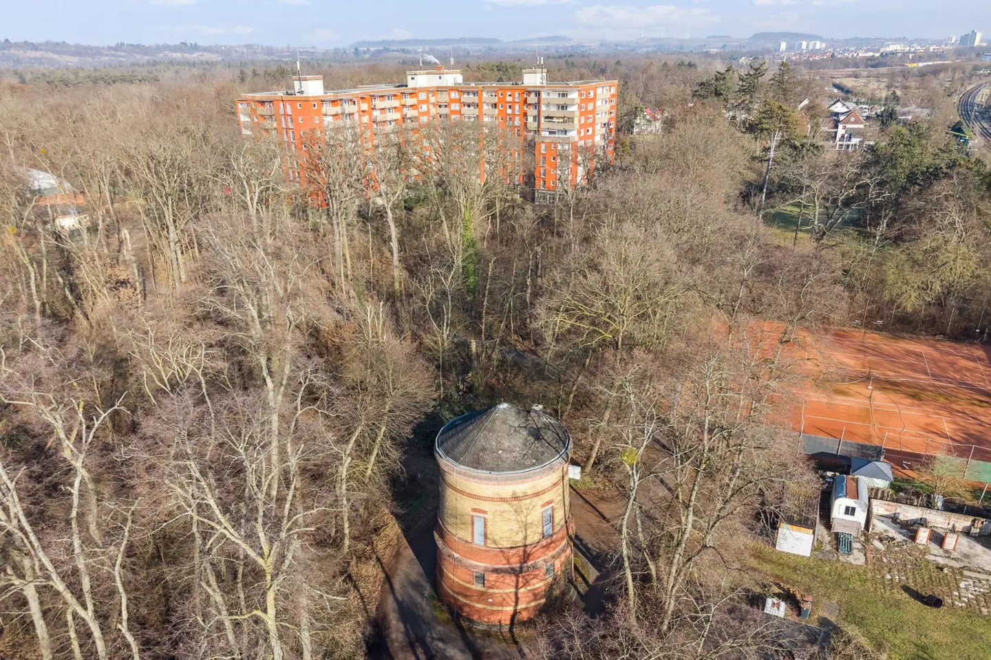 Aerial view of a brick water tower surrounded by bare trees, with an orange apartment building in the background.