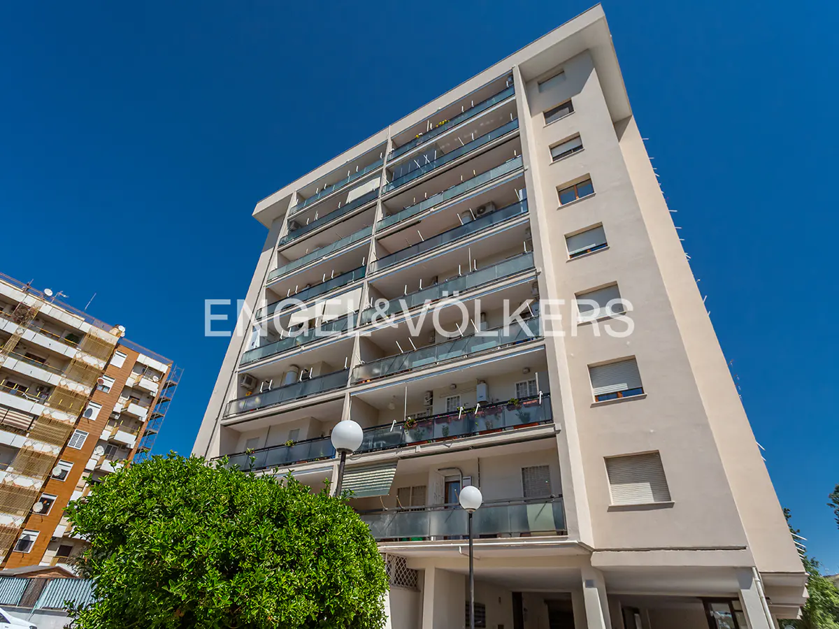 Exterior view of a tall, beige apartment building with glass balconies under a clear blue sky. Engel & Völkers logo visible.