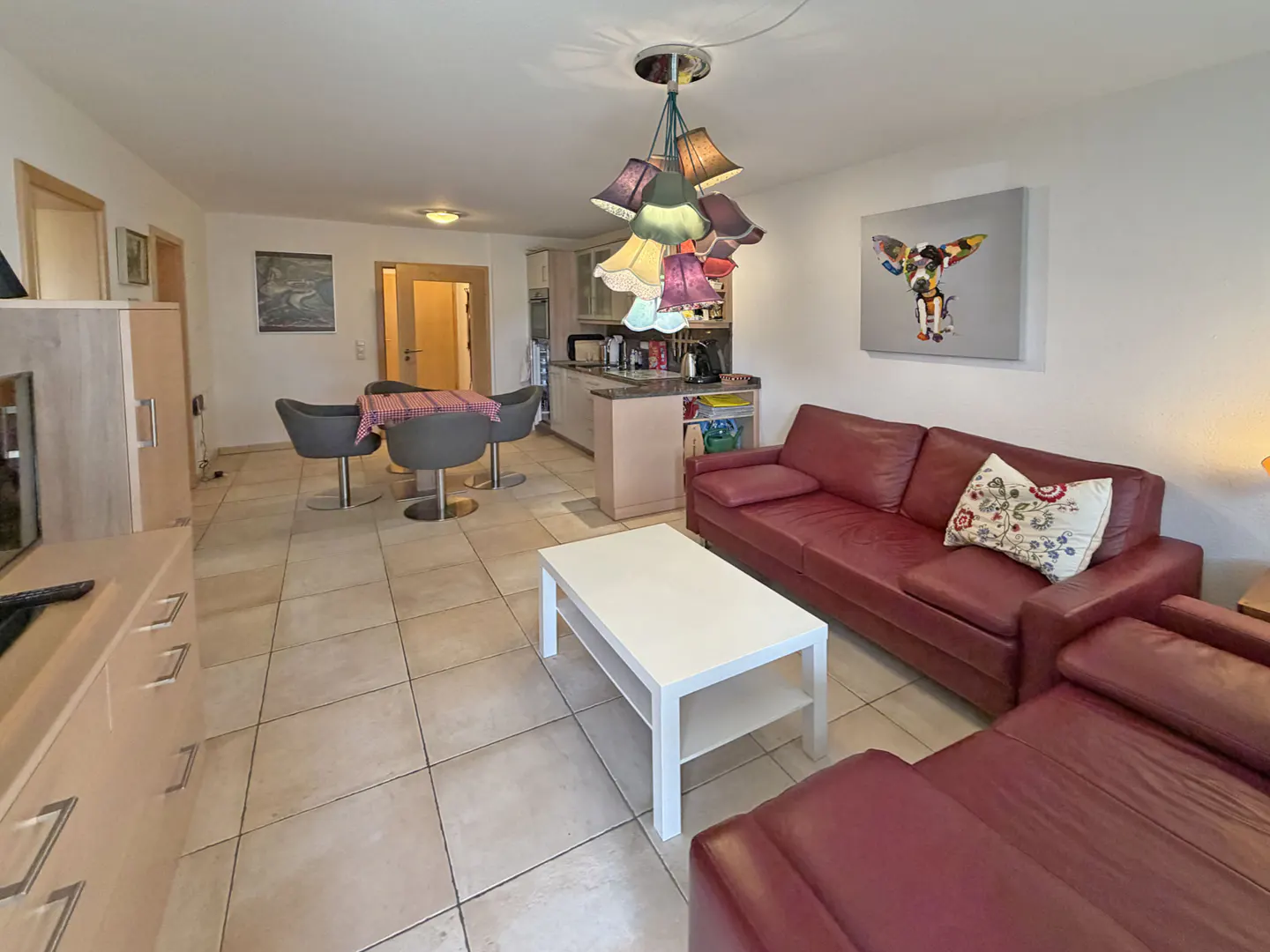 A living room with a red leather sofa, white coffee table, and a dining area with gray chairs. A colorful lampshade hangs above.