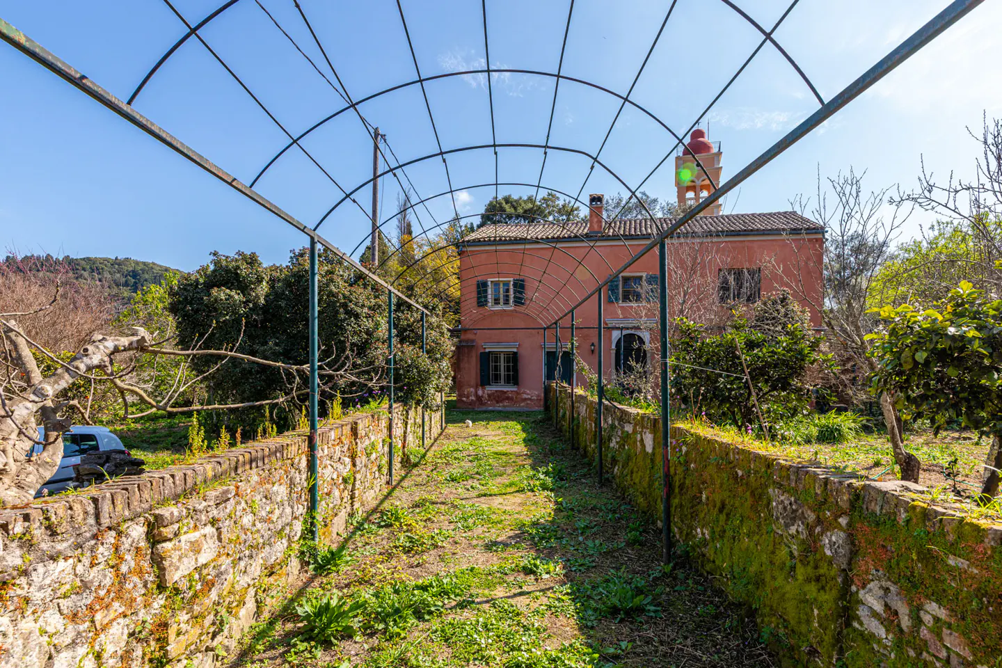 View of a salmon-colored house through a metal archway and mossy brick walls. A red-topped tower with a green light is visible.