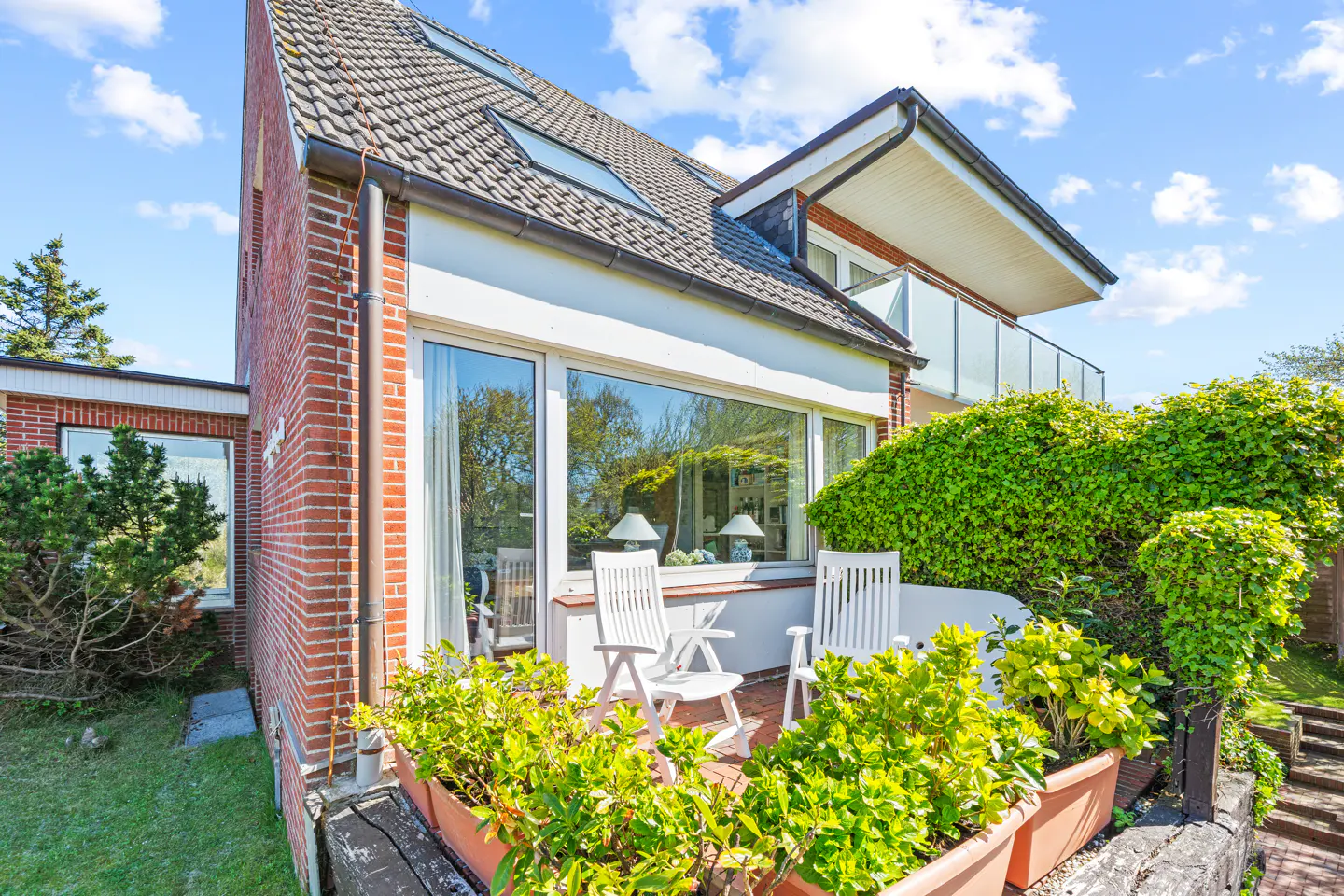 Exterior view of a two-story brick house with a tiled roof and a balcony. White chairs and plants adorn the patio.